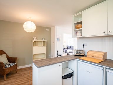 A kitchen area with white cabinets a wooden countertop a wicker chair and a small cupboard at Obo's Retreat in St Minver near Rock