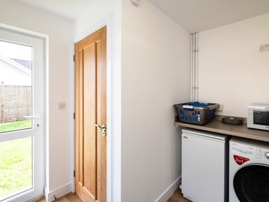 A utility room with a wooden door a glass door leading outside a washing machine a small fridge and a laundry basket on a countertop at Obos Retreat in St Minver near Rock