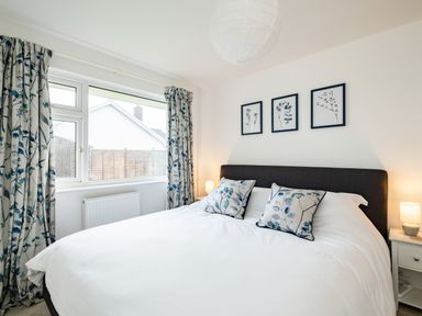 A bedroom with a large bed white bedding floral pillows two bedside tables with lamps and a window with floral curtains at Obos Retreat in St Minver near Rock