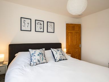 A bedroom with a bed with floral pillows wall art bedside tables with lamps and a wooden door at Obo's Retreat in St Minver near Rock