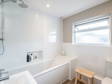 A bathroom with a bathtub shower combo a small wooden stool a window and a toilet at Obos Retreat in St Minver near Rock