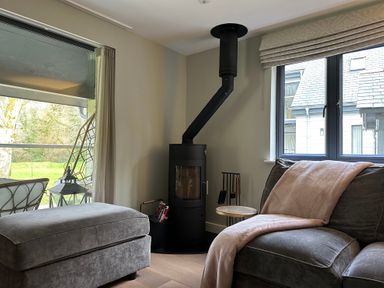 A living room with a wood stove next to a window and a gray armchair with a blanket at Primrose Hellandbridge near Bodmin