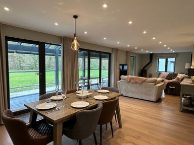 An open dining and living area with a wooden dining table set with plates and glasses and sofas near large windows at Primrose in Hellandbridge near Bodmin