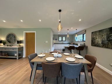 A dining room with a wooden table set with plates and glasses surrounded by chairs and an open kitchen with bar stools at Primrose Hellandbridge near Bodmin