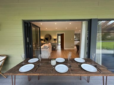 A wooden dining table set with plates and glasses on a covered patio looking into a living area at Primrose in Hellandbridge near Bodmin