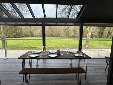 A dining table set with plates glasses and cutlery on a covered patio overlooking a grassy area and trees at Primrose in Hellandbridge near Bodmin