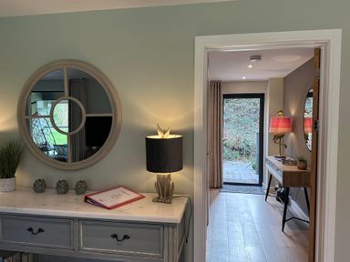 A hallway with a console table and lamp next to a round mirror leading to another room with a table lamp and door at Primrose Hellandbridge near Bodmin