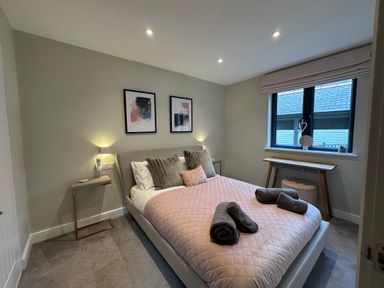 A bedroom with a double bed pink quilted bedspread and towels on it with bedside tables and framed pictures on the wall at Primrose Hellandbridge near Bodmin