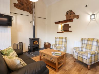 A living room with two plaid armchairs a black sofa a wooden coffee table a wood burning stove and a mounted television at Quoit X Barn in Stoodleigh near Bampton