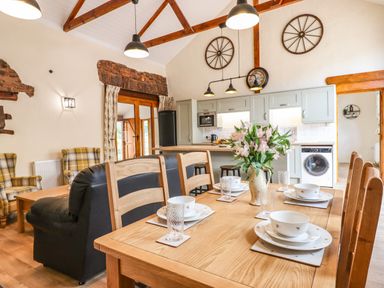A dining table set with bowls glasses and a vase with flowers in a kitchen and living room with wooden beams and stone accents at Quoit X Barn in Stoodleigh near Bampton