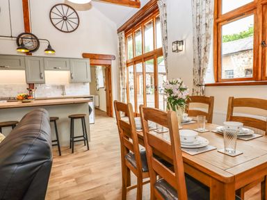 A dining area with a wooden table and chairs set for four next to large windows overlooking an outdoor stone building and a kitchen area with stools and cabinets at Quoit X Barn in Stoodleigh near Bampton