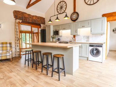 A kitchen with a wooden island counter and four stools a washing machine and a plaid armchair at Quoit X Barn in Stoodleigh near Bampton