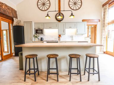 A kitchen with four wooden stools at a central island and wooden framed large windows at Quoit X Barn in Stoodleigh near Bampton