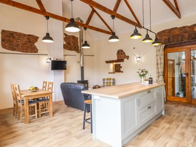 An open kitchen and dining area with a wooden dining table and chairs a kitchen island with hanging lights a sofa and wooden beams on the ceiling at Quoit X Barn in Stoodleigh near Bampton