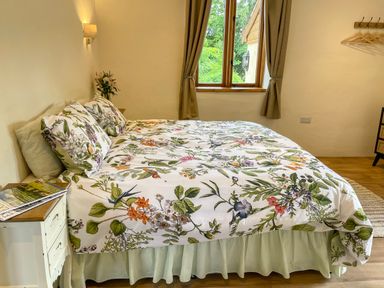 A bedroom with a bed with floral bedspread a window with curtains and a side table with books at Quoit X Barn Stoodleigh near Bampton