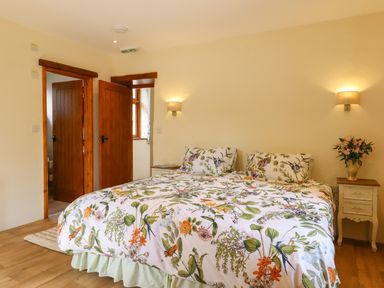 A bedroom with a double bed with floral bedding two wooden doors and bedside tables with lamps and flowers at Quoit X Barn in Stoodleigh near Bampton