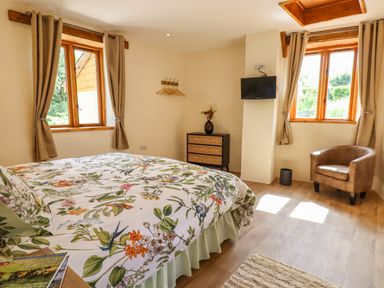 A bedroom with a floral bedspread two windows with curtains a small chest of drawers a wall-mounted tv and a brown armchair at Quoit X Barn Stoodleigh near Bampton