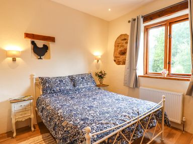 A bedroom with a bed covered in blue floral bedding beside bedside tables with flowers and magazines at Quoit X Barn in Stoodleigh near Bampton