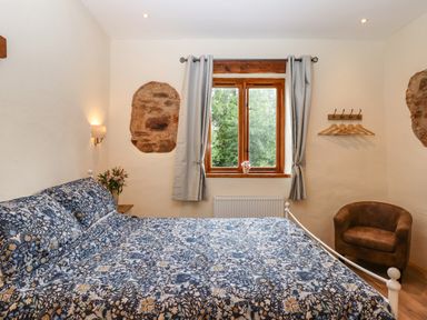 A bedroom with a bed covered with a floral patterned duvet next to a window with gray curtains and a brown armchair at Quoit X Barn in Stoodleigh near Bampton