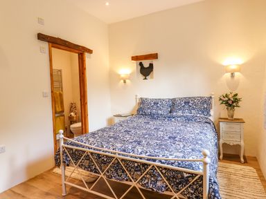 A bedroom with a double bed in blue floral bedding two bedside tables with flowers and a toilet visible through a wooden framed door at Quoit X Barn in Stoodleigh near Bampton