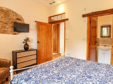 A bedroom with a patterned bedspread a wooden chest of drawers a mounted television and an open door to a bathroom at Quoit X Barn in Stoodleigh near Bampton