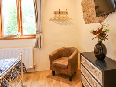 A corner of a bedroom with a brown armchair beside a small dresser with a vase and flowers and a wall-mounted TV at Quoit X Barn in Stoodleigh near Bampton
