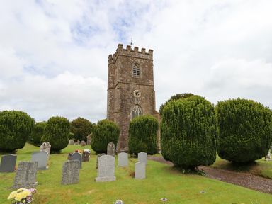 A church with a clock tower surrounded by gravestones and trimmed trees at Quoit X Barn in Stoodleigh near Bampton