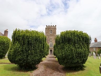 A stone church with a clock tower behind large trimmed bushes and a gravel path at Quoit X Barn in Stoodleigh near Bampton