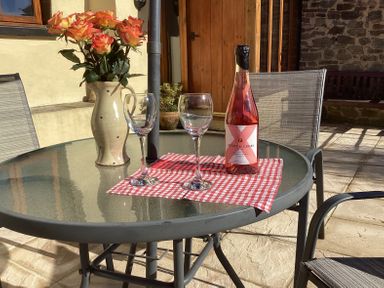 A glass patio table with two wine glasses a bottle of rose wine and a vase of roses with chairs around on a stone patio at Quoit X Barn in Stoodleigh near Bampton
