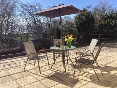 An outdoor patio with a round glass table under a large umbrella and four chairs with flowers and coffee cups on the table at Quoit X Barn in Stoodleigh near Bampton