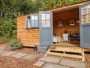 A cabin with open double doors and a window showing wooden interior with furniture and plants outside at Hideaway Cabin in Cadbury near Thorverton