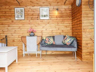 A wooden room with a gray sofa with patterned cushions a small table with flowers and two framed pictures on the wall at Hideaway Cabin in Cadbury near Thorverton