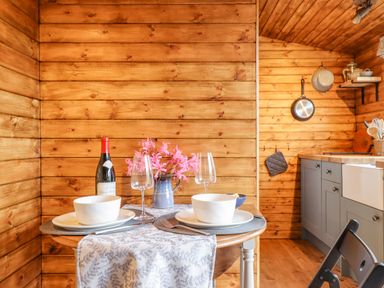 A small dining table set for two with bowls glasses wine bottle and flowers in a wooden kitchen at Hideaway Cabin in Cadbury near Thorverton