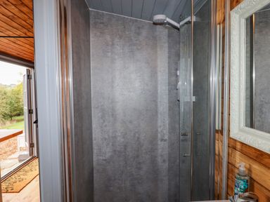 A bathroom shower area with glass door a mirror bottle of hand soap and an open door showing an outside view at Hideaway Cabin in Cadbury near Thorverton