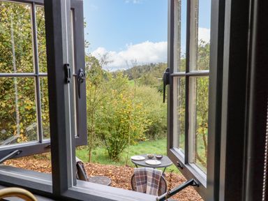 An open window looking out onto a small round table with plates and bowls on a patio surrounded by trees at Hideaway Cabin in Cadbury near Thorverton
