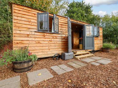 A wooden cabin with open doors and windows surrounded by plants and stone pathway at Hideaway Cabin in Cadbury near Thorverton