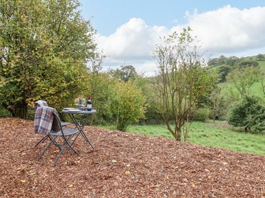 A small table with two chairs and a bottle of wine on a wood chip ground surrounded by trees and grass at Hideaway Cabin in Cadbury near Thorverton