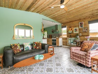 A living room with a black sofa and colorful cushions a patterned armchair a wooden coffee table and a kitchen area in the background at Lulu Logs in Congresbury
