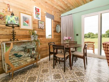 A dining area with a round wooden table and four chairs beside sliding glass doors and a glass cabinet against a wooden wall at Lulu Logs in Congresbury