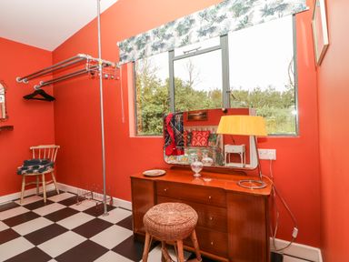 A dressing room with a wooden dresser a round wicker stool a yellow lamp and a chair with folded blankets at Lulu Logs in Congresbury