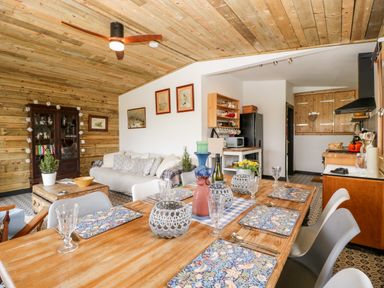 A dining table set with placemats and glassware in a room with a wooden ceiling and kitchen in the background at Down the Rabbit Hole in Brinsea near Congresbury