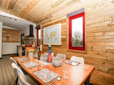 A dining table set for six with placemats and glassware in a wooden paneled room with a kitchen in the background at Down the Rabbit Hole in Brinsea near Congresbury