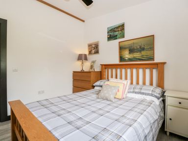A bedroom with a wooden bed frame and plaid bedding with a chest of drawers and lamp at Down the Rabbit Hole Brinsea near Congresbury