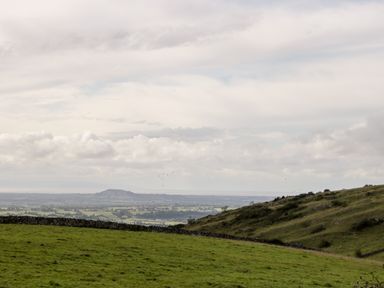 A grassy hillside with a stone wall and distant hills under a cloudy sky at Down the Rabbit Hole in Brinsea near Congresbury