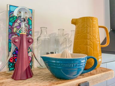 A wooden shelf with a Le Creuset blue citrus juicer, a yellow pitcher, three clear bottles, and a red corkscrew in front of a stained glass panel at Down the Rabbit Hole in Brinsea near Congresbury