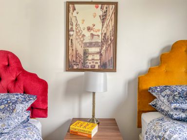 A bedroom with a bedside table holding a lamp and two books between a red and a yellow bed at Down the Rabbit Hole in Brinsea near Congresbury