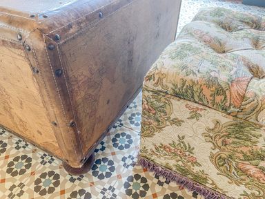 A close-up of a leather trunk and a floral fabric ottoman on a patterned tile floor at Down the Rabbit Hole in Brinsea near Congresbury