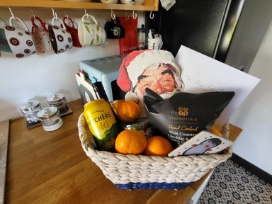 A kitchen counter with mugs hanging above and a gift basket containing fruit snacks a can and a Santa decoration at Down the Rabbit Hole in Brinsea near Congresbury