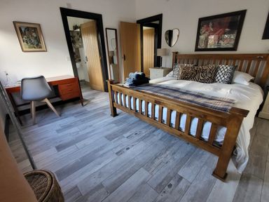 A bedroom with wooden bed, desk with chair, nightstand with lamp, and framed pictures on walls at Down the Rabbit Hole in Brinsea near Congresbury