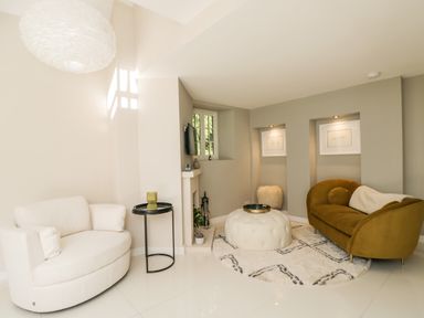 A living room with a white rounded armchair a small black side table a beige round ottoman a brown sofa and framed art on walls at The Hayloft in Sherston
